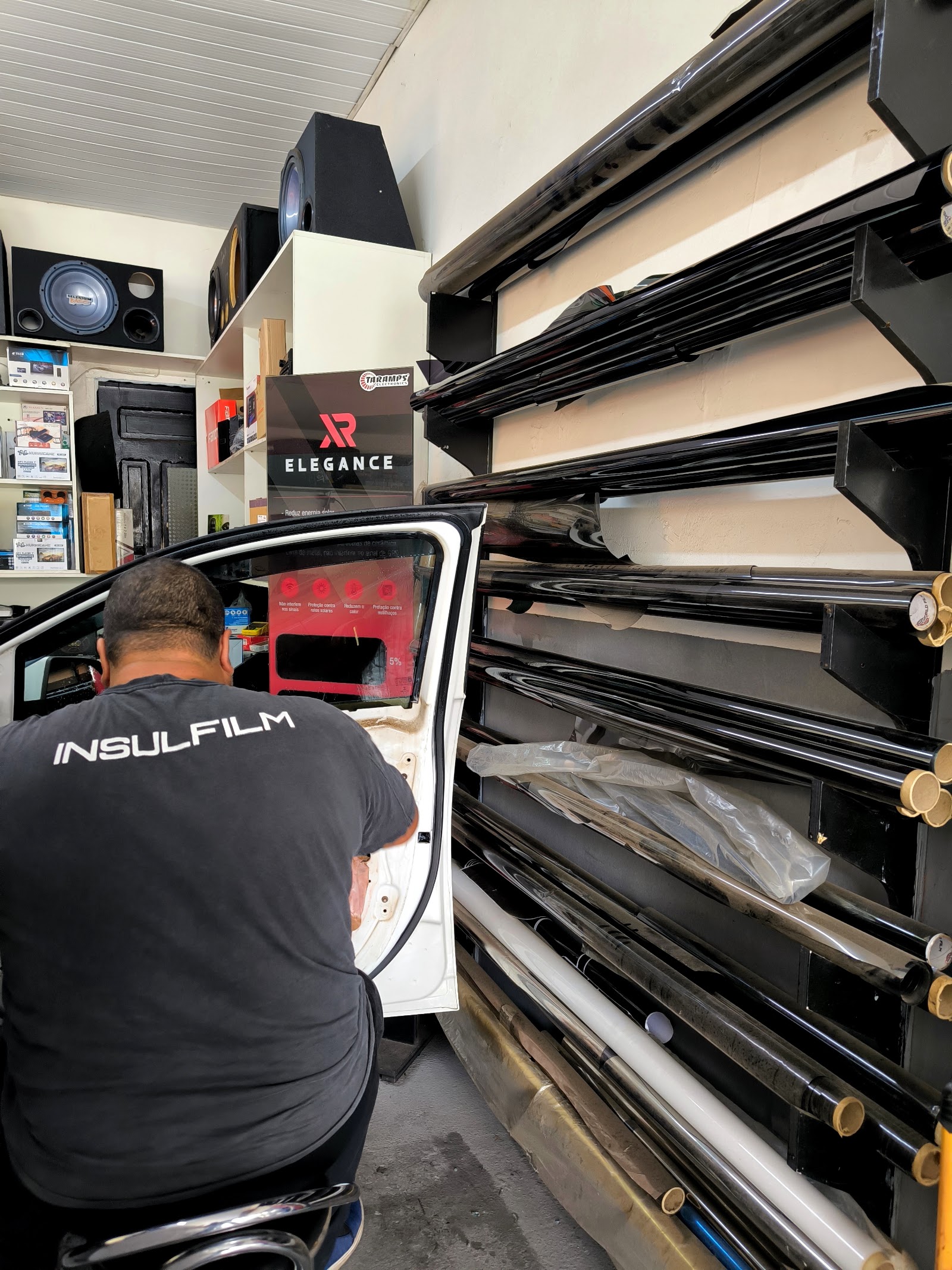 Profissional da World Sound 1, de costas, aplicando película na porta de um carro, com bobinas de insulfilm na parede ao lado