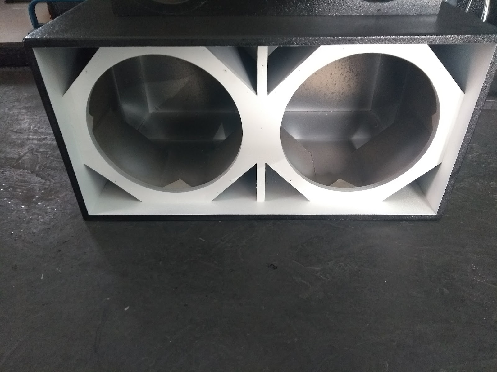 Caixa dutada preta com interior branco e dois recortes para subwoofers, apoiada no chão da loja